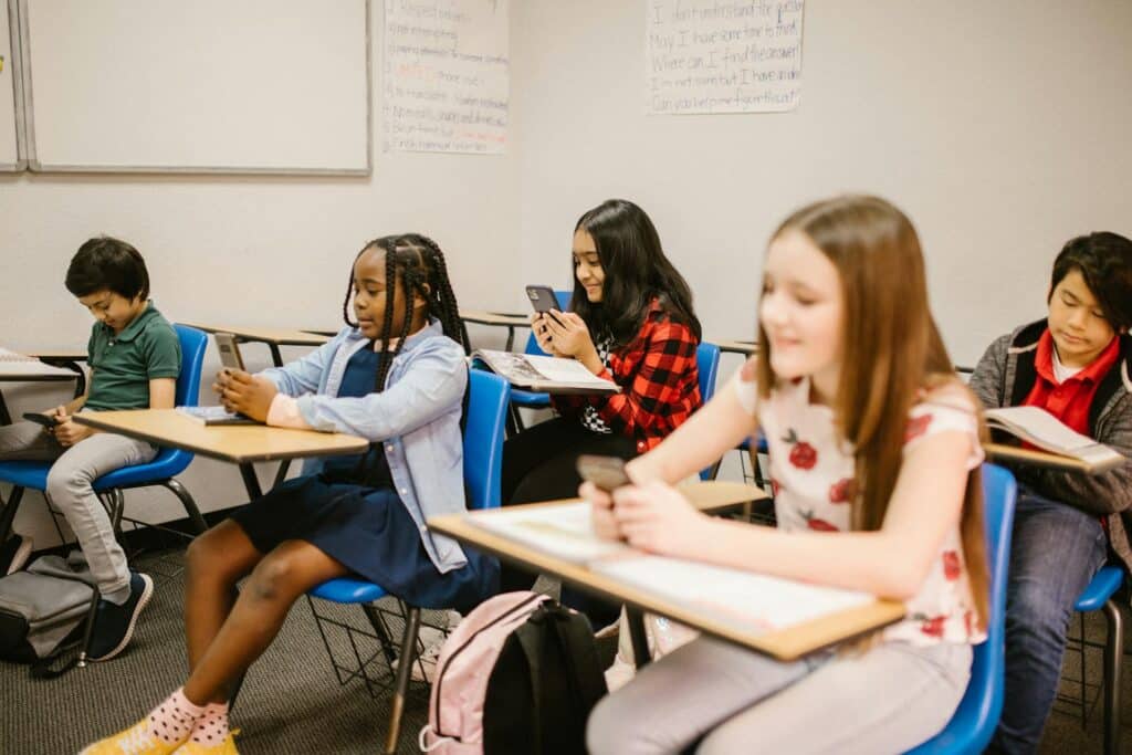 Kinder im Klassenzimmer, sitzend auf ihrem Schreibtisch, schauen auf ihr Handy