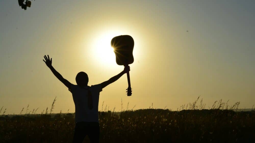 Mensch steht auf einem Feld draußen, mit Hände gespreizt nach oben, an einer Hand hält die Person eine Gitarre, sonniger Himmel